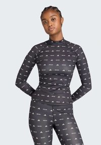 Γυναίκα με πλεξούδες φοράει εφαρμοστό μαύρο μακρυμάνικο μπλουζάκι και κολάν με λευκά σχέδια "MCMN GECT" και λογότυπα, στέκεται με τα χέρια πίσω από την πλάτη.