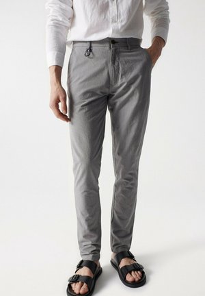 Mężczyzna w dopasowanych szarych spodniach, białej koszuli z długim rękawem, czarnych sandałach z klamrą, z jedną ręką w kieszeni i karabińczykiem przypiętym do paska.