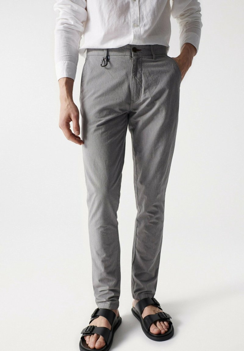 Homme portant un pantalon gris ajusté, une chemise blanche à manches longues, des sandales noires à boucle, avec une main dans la poche et un mousqueton accroché à la ceinture.