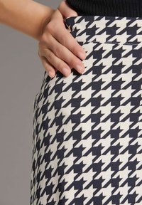 Hoch geschnittenen Rock mit einem schwarz-weißen Hahnentritt-Muster, aus einem glatten Stoff gefertigt. Eine Hand liegt auf dem Bund für besseren Halt.
