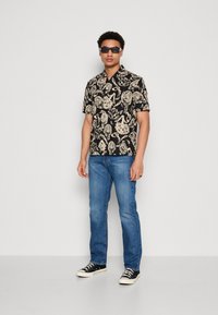 Schwarzes kurzärmliges Shirt mit einem beigen Blumenmuster, hellblaue Jeans und schwarze Turnschuhe, getragen mit einer dunklen Sonnenbrille.