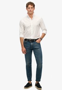 Camisa blanca de botones, jeans azul ajustados, cinturón negro y zapatos negros de tipo slip-on. El modelo está de pie frente a un fondo blanco liso.