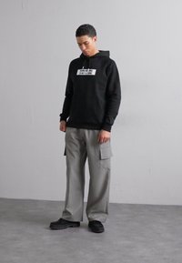 Sweatshirt à capuche noir avec texte sur le devant, associé à un pantalon cargo gris clair avec des poches latérales et des chaussures noires.