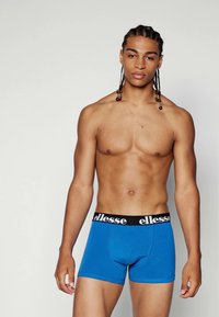 Blaue Baumwoll-Boxershorts mit einem schwarzen elastischen Bund, der das "ellesse"-Logo trägt. Der Stoff wirkt glatt und enganliegend.
