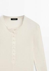 Chemise henley beige à manches longues avec sept boutons sur le devant et un col rond, étiquetée Massimo Dutti.