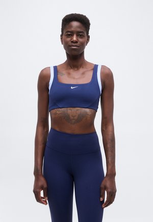 Αθλητική γυναίκα με κοντά μαλλιά φοράει σκούρο μπλε αθλητικό σουτιέν και κολάν, δείχνοντας ένα τατουάζ λιονταριού στο πάνω μέρος της κοιλιάς της.