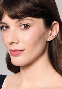 Srebrne kolczyki w kształcie sztyftów z pojedynczym przejrzystym kryształem, noszone przez modelkę z ciemnymi włosami w naturalnym makijażu.