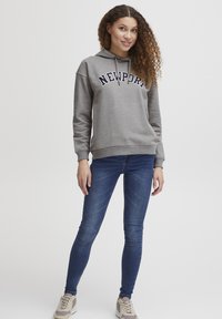 Felpa grigia con testo "NEWPORT" in blu navy, tasca a canguro e cappuccio con drawstring. Abbinata a jeans skinny blu scuro e sneakers beige.