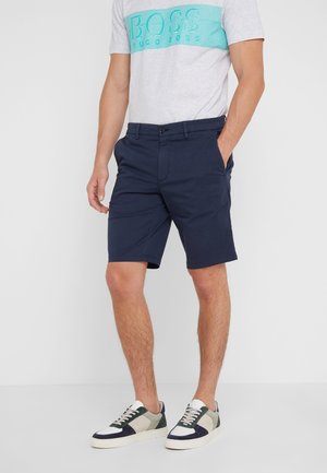 Uomo con pantaloncini blu navy, maglietta grigia e turchese con logo BOSS e sneakers bianche con dettagli neri e verdi, in piedi con una mano in tasca.