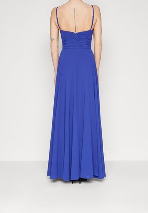 Femme portant une robe bleu roi longue jusqu'au sol avec de fines bretelles et un dos froncé, debout devant un fond clair uni.