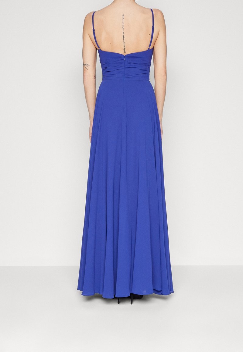 Femme portant une robe bleu roi longue jusqu'au sol avec de fines bretelles et un dos froncé, debout devant un fond clair uni.