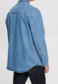 Světle modrá denimová košile s oblou hemem, dlouhými rukávy a límcem na knoflíky. Jsou viditelné švy a detail zadního dílu.