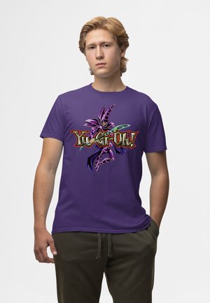 Junger Mann mit hellem Haar, der ein lila T-Shirt mit der Dark Magician-Figur und dem Yu-Gi-Oh!-Logo trägt, steht mit einer Hand in der Tasche.