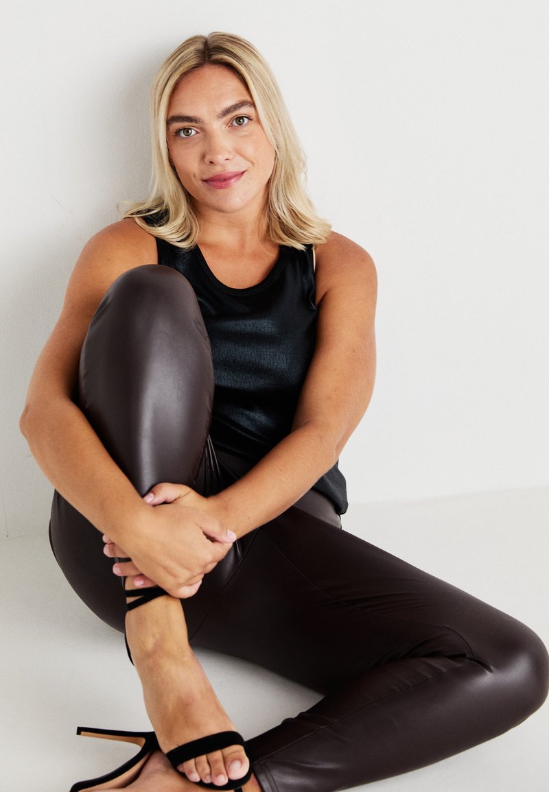 Black sleeveless top paired with shiny, fitted brown leggings. Model sits with legs crossed, wearing black strappy high heels. White background.