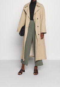 Beige pitkä takki leveillä läpillä, yhdistettynä oliivinvihreisiin leveälahkeisiin housuihin. Musta kapea poolopaita ja nauhalla varustetut mustat sandaalit viimeistelevät asun.