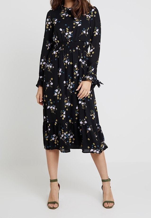 Femme portant une robe midi noire à manches longues avec motif floral, taille cintrée et ourlet volanté, associée à des talons olive à bride cheville.