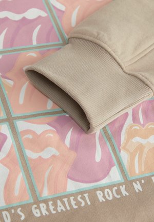 Beige sweatshirtmanchet met ribbelstructuur, rustend op een kleurrijke grafische achtergrond met lippen en tekst in pastelkleuren.