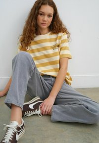 Geel en crème gestreepte t-shirt met de tekst "Portofino", gecombineerd met lichtgrijze baggy broeken en bruine Nike sneakers, zittend op de vloer.