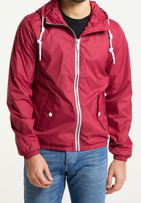 Chaqueta impermeable roja con cremallera frontal, puños elásticos, capucha ajustable, dos bolsillos laterales y una textura lisa y brillante.