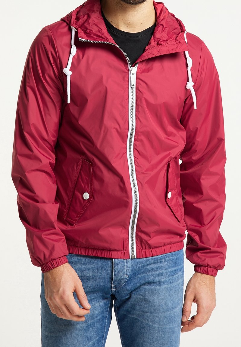 Chaqueta impermeable roja con cremallera frontal, puños elásticos, capucha ajustable, dos bolsillos laterales y una textura lisa y brillante.