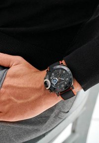 Orologio cronografo nero con accenti arancioni, cinturino testurizzato, grandi numeri, sottocompiti e una cassa resistente. Indossato su un polso contro un indumento scuro.