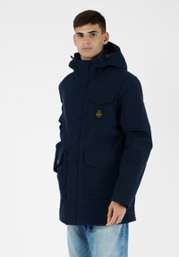 Parka con cappuccio di colore navy, dotata di molteplici tasche frontali, chiusura con zip e un emblema testurizzato sul petto. Indossata con jeans azzurri chiari.