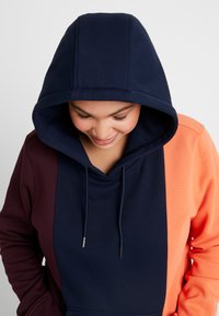 Sudadera con capucha que presenta paneles en navy, burdeos y naranja, con una textura suave y detalles metálicos en los cordones de la capucha.