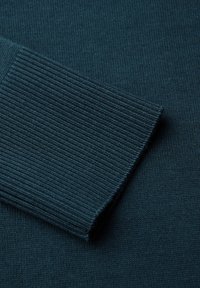 Türkis gestrickter Stoff mit einem gerippten Bündchen. Glatte Textur mit feinen Stickdetails. Nahaufnahme, die die Dehnbarkeit und Weichheit des Stoffs zeigt.