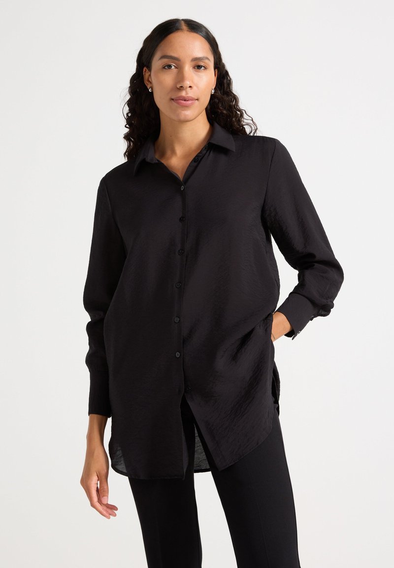 Camicia nera a maniche lunghe realizzata in tessuto leggero e testurizzato, con chiusura a bottoni frontale, colletto e spacchi laterali per facilitare il movimento.