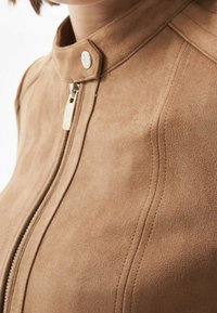 Béžová semišová bunda se stojáčkem, stříbrným zipem a viditelnými švy, s hladkou texturou a na míru střiženým tvarem.