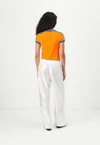 Vrouw met lang, krullend haar, gekleed in een oranje t-shirt met blauwe rand en losse witte satijnen broek, staand tegen een effen witte achtergrond.