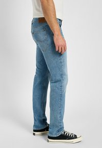 Ljusblå denimjeans med rak passform, med en brun läderlapp och standard femfickorsdesign, kombinerat med svarta segeltossor.