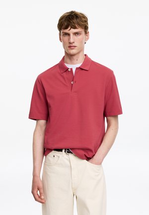 Jeune homme aux cheveux châtain clair portant un polo rouge à manches courtes et un pantalon écru, debout avec une main dans la poche.