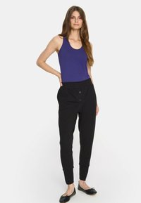 Kobieta ubrana w bezrękawnik w kolorze purpurowym, czarne spodnie z wysokim stanem z guzikami oraz czarne baletki, stoi z jedną ręką na biodrze.