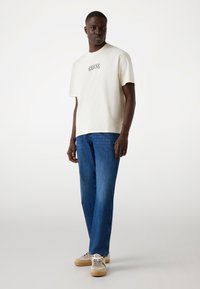 Camiseta corta de manga corta color crema con un logo negro de "GUESS", combinada con unos jeans rectos azules y zapatillas beige con cordones blancos.