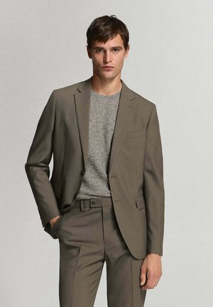 Joven con un traje marrón y un jersey de punto gris, de pie con una mano en el bolsillo frente a un fondo claro y liso.