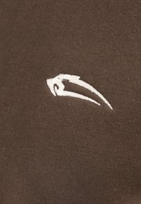 Tela marrón con una textura suave que presenta un logo bordado en blanco en el lado izquierdo. El diseño es simple y minimalista.