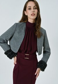 Giacca grigia corta con polsini in pelliccia sintetica nera, abbinata a una blusa bordeaux e una gonna con dettaglio in oro sulla vita.