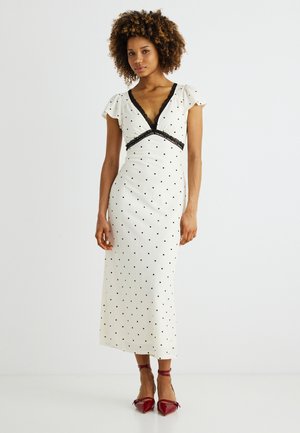 Frau mit lockigem Haar, die ein weißes Midikleid mit schwarzen Polka-Dots und Spitzenbesatz trägt, kombiniert mit roten Knöchelriemen-Absätzen, stehend vor weißem Hintergrund.