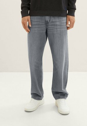 Man wearing light blue straight-leg jeans, black long-sleeve top, and white sneakers standing on a light floor.