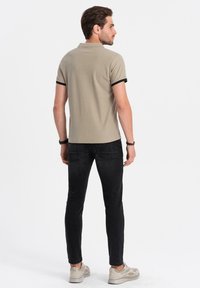 Beige polo tričko s černými detaily, přiléhavý střih. Nosené s úzkými černými džínami a světlými teniskami. Zobrazeno zezadu, s minimálními detaily.
