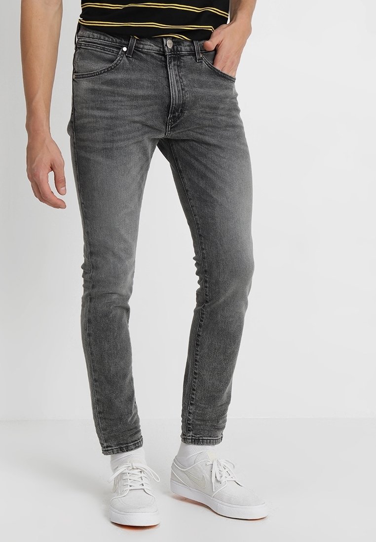 Mann trägt ausgewaschene schwarze Slim-Fit-Jeans, weiße Socken, weiße Nike-Sneaker und ein schwarzes Hemd mit gelben Streifen.
