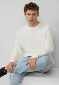 Helles graues Rundhals-Sweatshirt mit langen Ärmeln und strukturierter Stoff, kombiniert mit hellblauen Jeans und schwarzen Turnschuhen. Einfaches Design.