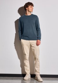 Mörkblå sweatshirt med rund halsringning, långa ärmar, strukturerad känsla och en liten logotyp. Matcher med ljusbeiga byxor och ljusa sneakers.