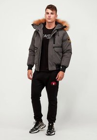 Casaco cinzento acolchoado com capuz forrado a pelo, fecho de correr com bolsos laterais, vestido sobre uma camisa preta e calças de treino pretas a condizer, combinado com ténis.