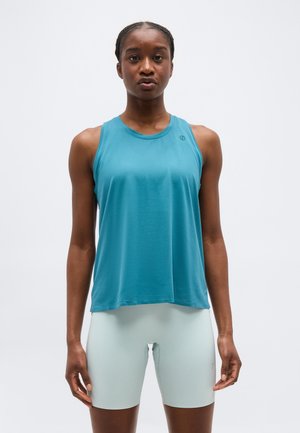 Mujer de pie de frente, vestida con una camiseta deportiva sin mangas de color verde azulado y unos pantalones cortos de ciclismo ajustados de color gris claro, sobre un fondo liso.