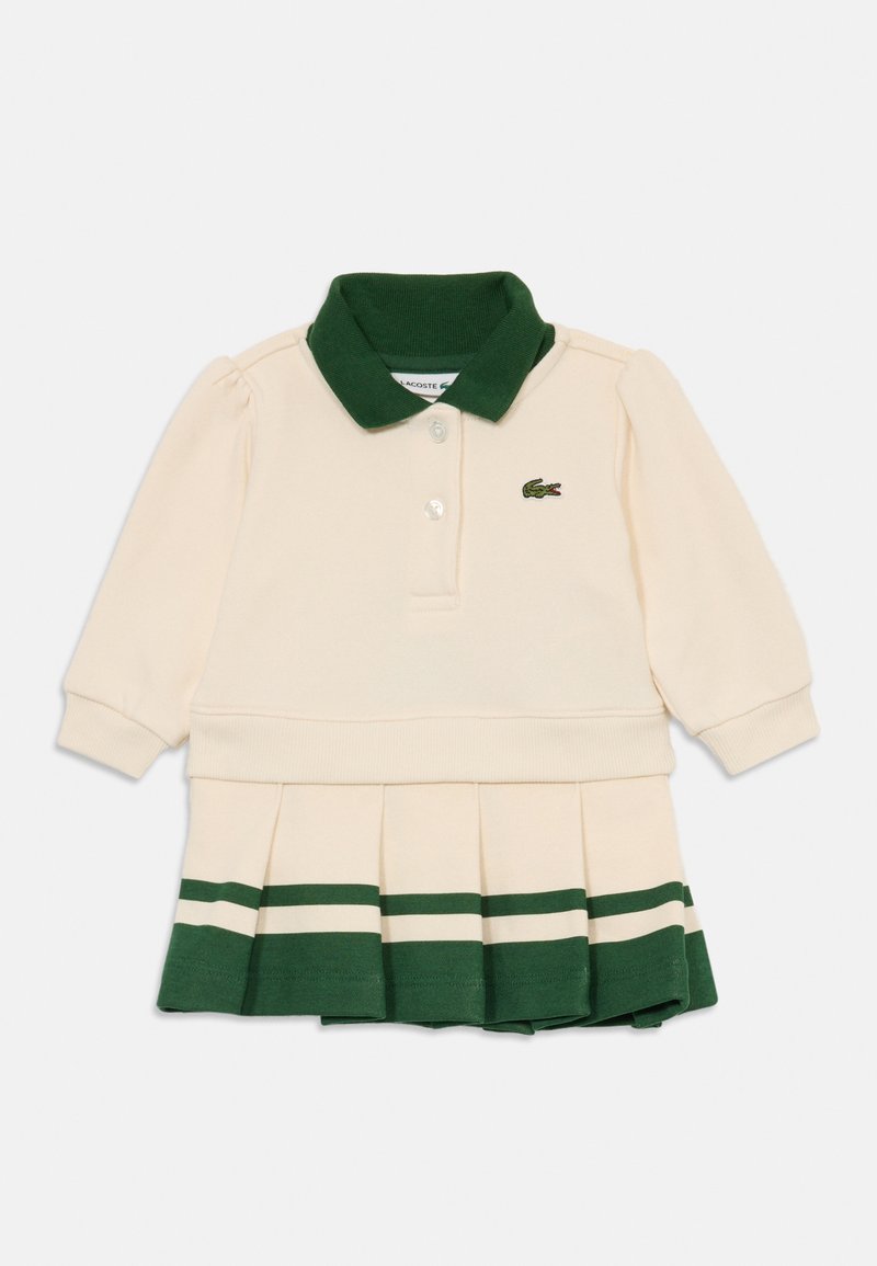 Kremfarget langermet pologenserkjole med grønn krage, to knapper, plisserte skjørt med grønne og kremfargede striper, og liten logo på brystet.