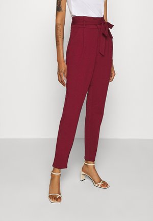 Persoon draagt bordeauxrode tapered broek met hoge taille, een strikceintuur en witte sandaal met hak en bandjes, staand tegen een effen achtergrond.