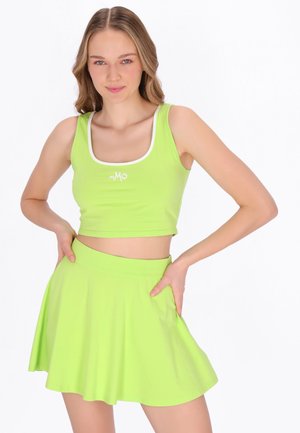 Fiatal nő lime zöld, rövid felsőt és hozzáillő kitágított szoknyát visel, kezét a csípőjén tartva pózol fehér háttér előtt.