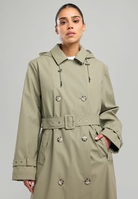 Khaki säänkestävä trench coat, jossa on kaksoisnappi-malli, huppu, vyötäröä korostava vyö ja suuret napit. Sileä pinta.